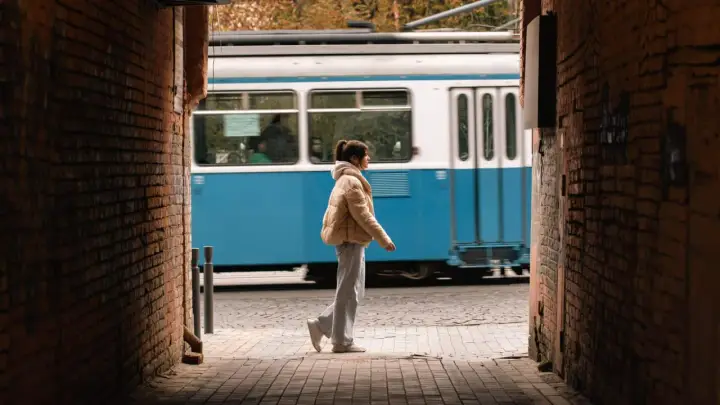 Una ragazza cammina in un vicolo, un tram blu sfreccia sullo sfondo. Una foto vedo non vedo che cattura un momento fugace.