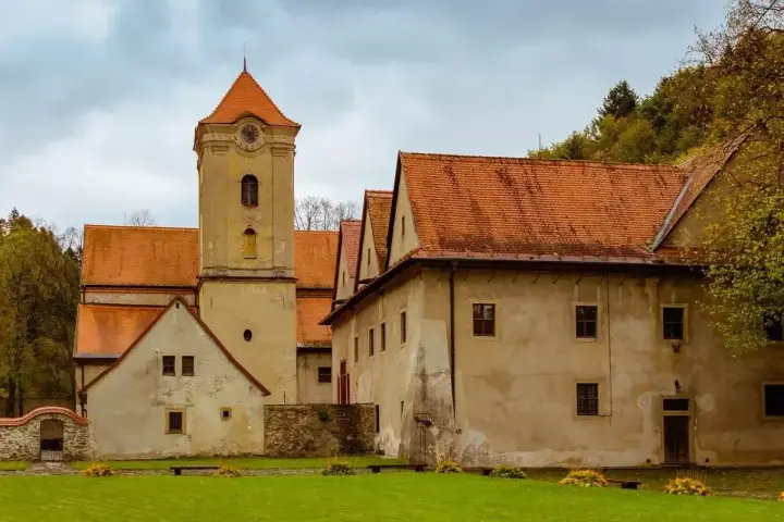 Czerwony klasztor w Pieninach, z charakterystyczną wieżą zegarową i pomarańczowymi dachami, otoczony zielonym trawnikiem i drzewami.