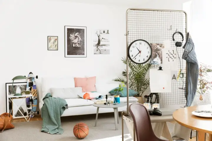 En avslappnad inredning hemma med vit soffa, konst på väggen, en basketboll och en skateboard. En stor klocka hänger på ett metallgaller.