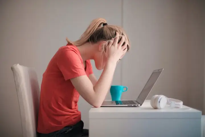 Kobieta z rękami na głowie przy laptopie, kubek i słuchawki. Czy to objawy wypalenia zawodowego jak sobie radzić?