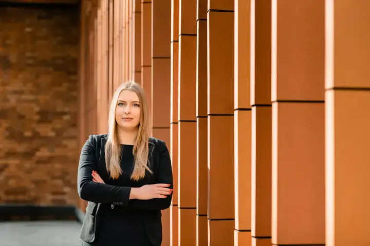 Kobieta w czarnym garniturze stoi na tle ceglanej ściany i pomarańczowych pionowych elementów.