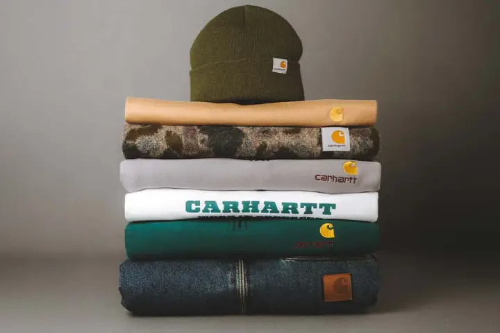 Stos ubrań Carhartt: czapka, bluzy i jeansy. Carhartt to firma znana z wytrzymałej odzieży roboczej i streetwearowej.