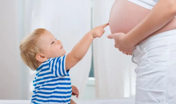 Mały chłopiec w pasiastej koszulce dotyka paluszkiem brzucha mamy, która jest w 25 tygodniu ciąży.