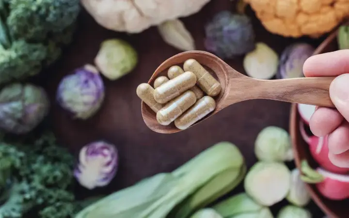 Łyżka z kapsułkami – witaminy na energię. W tle świeże warzywa: brokuły, brukselka, kapusta pak choi.
