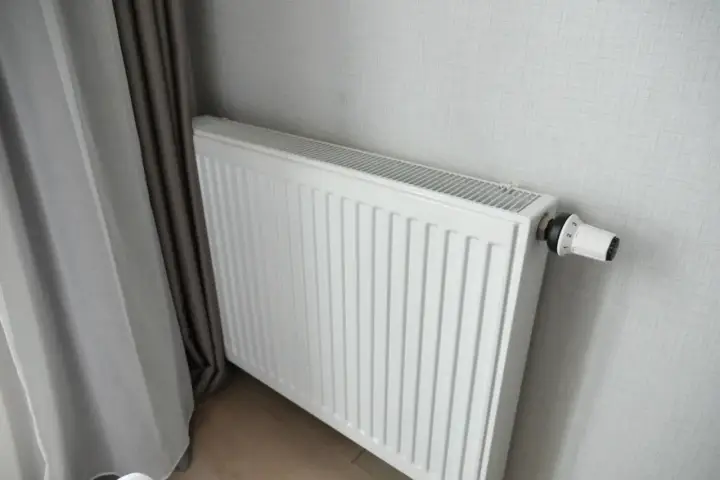 Biały grzejnik panelowy z termostatem, idealny do pokoju 16m2. Obok zasłony.