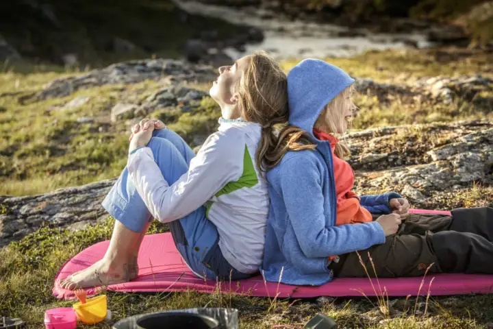 Två personer, en vuxen och ett barn, sitter rygg mot rygg på en liggunderlag i naturen. En härlig stund att vandra med barn.