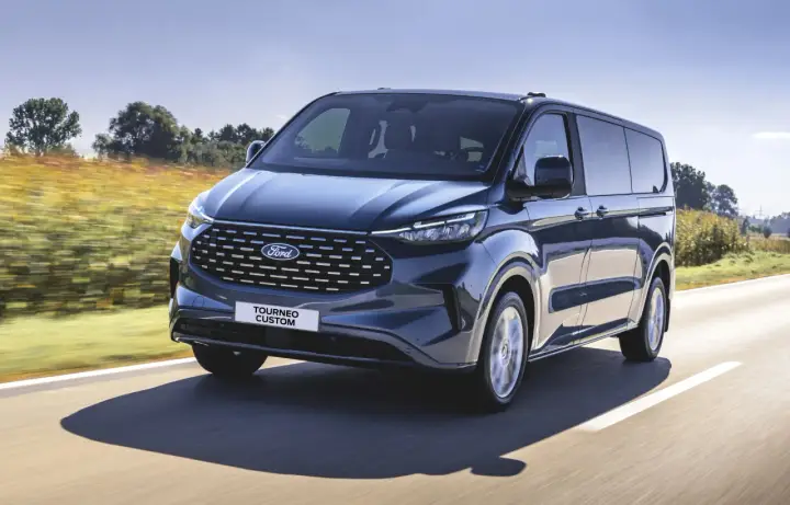 Ciemnoniebieski ford 7 osobowy Tourneo Custom pędzi po drodze wśród pól.