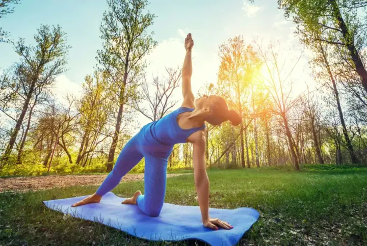 Kvinna utför yoga utomhus i en skogsmiljö. Solen skiner genom träden.