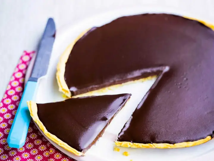 Une part de la meilleure recette de tarte au chocolat, avec une ganache brillante sur une pâte dorée. Un couteau bleu est à côté.