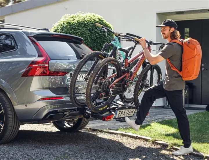 Mężczyzna z pomarańczowym plecakiem montuje bagażnik rowerowy na hak do Volvo XC60. Dwa rowery górskie są już zamocowane. Przepisy dotyczące bagażnika rowerowego na hak są kluczowe dla bezpieczeństwa.