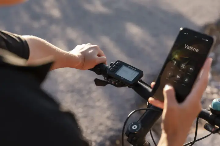 Rowerzysta trzyma telefon, na którym wyświetla się połączenie z Valerie. Obok widoczne są symbole na liczniku rowerowym, pokazujące prędkość i inne dane.