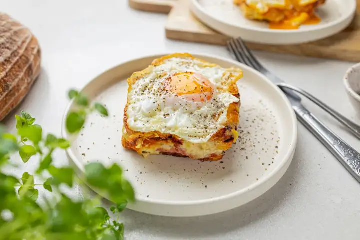 Pyszny croque madame z jajkiem sadzonym na wierzchu, na talerzu, obok chleba i zielonych liści.