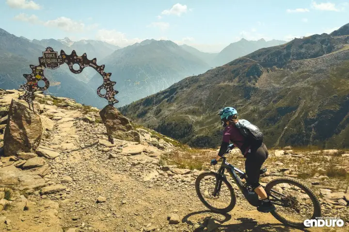 Kobieta na rowerze zjeżdża po kamienistym szlaku w bike parku, pod bramą z napisem "Bike Republic Solden".