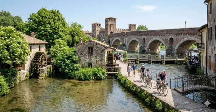 Un pittoresco borghetto sul Mincio con mulini ad acqua, un ponte ad archi e persone che passeggiano e pedalano.