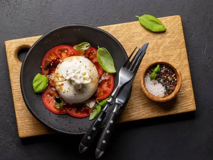 Pyszna burrata z pomidorami, polana oliwą, udekorowana bazylią. Obok miseczka z solą i pieprzem.
