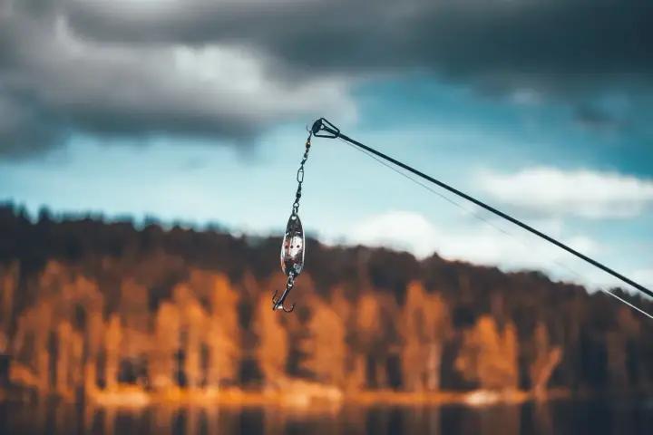 Przynęta z haczykiem wisi na żyłce, gotowa do połowu na jeziorze Skarlińskim. W tle jesienne drzewa i błękitne niebo.