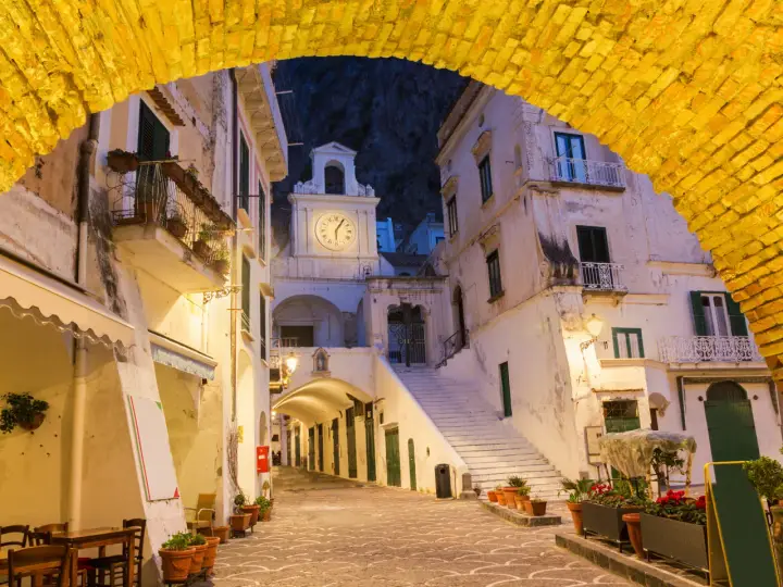 Un vicolo illuminato ad Atrani, con un arco giallo in primo piano e un campanile con orologio sullo sfondo.