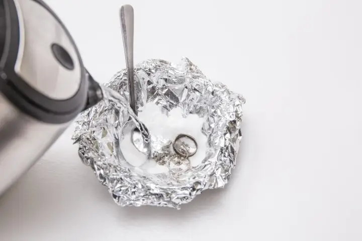 Nettoyage d'argenterie noircie : de l'eau chaude est versée sur des bijoux en argent et une cuillère dans une coupelle en papier aluminium.