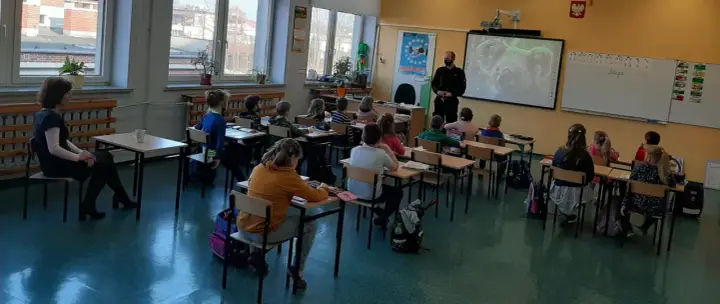 Dzieci słuchają lekcji o bezpieczeństwie w szkole. Nauczycielka i policjant prowadzą zajęcia.