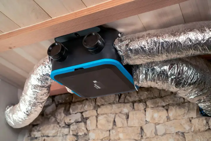 Unité de ventilation maison avec conduits argentés, installée sous un plafond en bois et au-dessus d'un mur en pierre.
