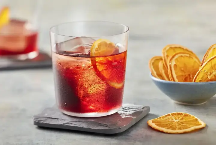 Klasyczny negroni przepis: czerwony koktajl z lodem i plasterkiem pomarańczy, obok miseczka suszonych pomarańczy.