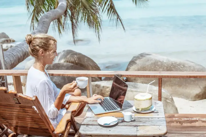 Kvinna med kaffe och laptop på en strandterrass, drömmen om ett digitalt nomad jobb.