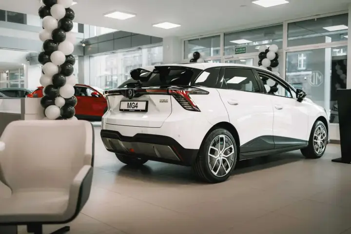 Białe auto MG 4 Electric w salonie, ozdobionym balonami. Obok czerwony samochód i wygodne fotele.