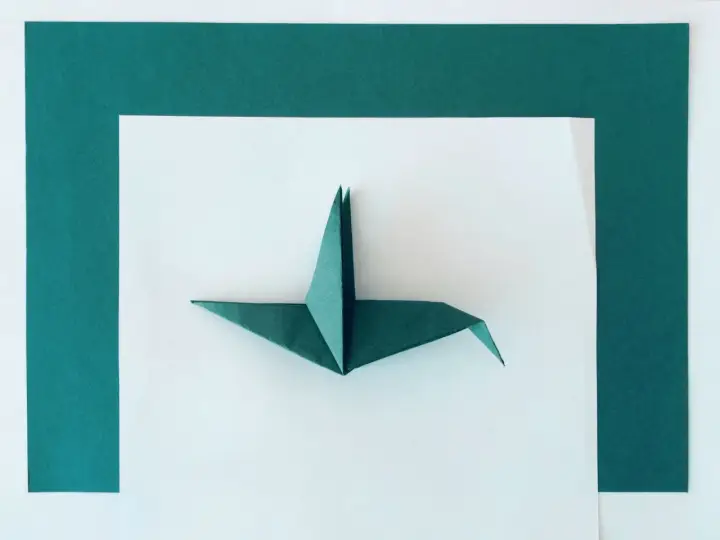 Zielony żuraw z papieru, przykład sztuki origami dla dorosłych, leży na białym tle otoczonym ciemnozielonymi ramkami.