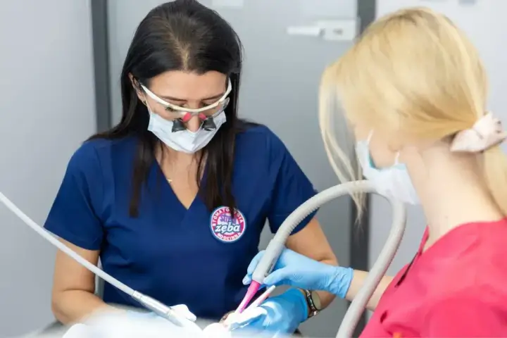 Dwie dentystki w maseczkach i okularach powiększających pracują przy pacjencie. Jedna używa wiertła, druga asystuje, podając anestetyki.