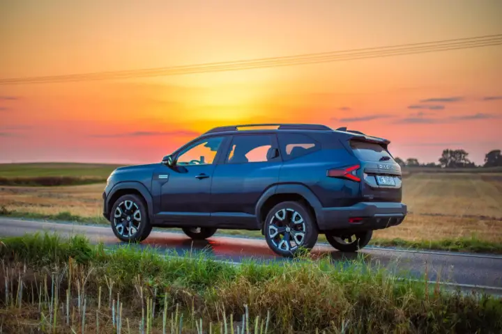 Ciemnoniebieski Dacia Bigster 2025 na tle zachodzącego słońca, na wiejskiej drodze.