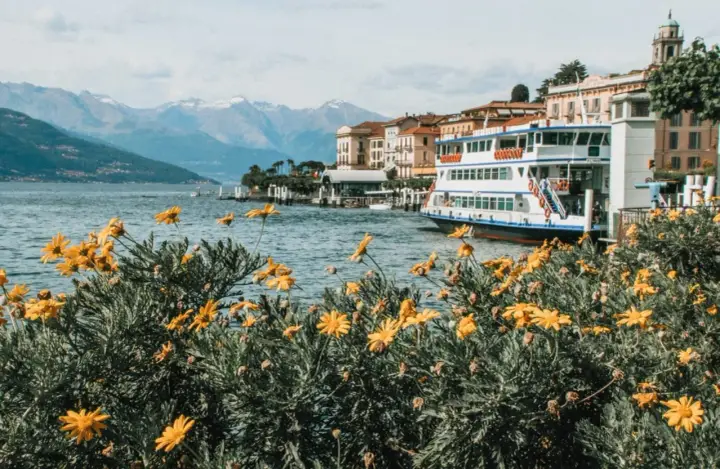 Fiori gialli in primo piano, lago blu, montagne innevate e un battello attraccato. Una perfetta gita fuori porta Milano.