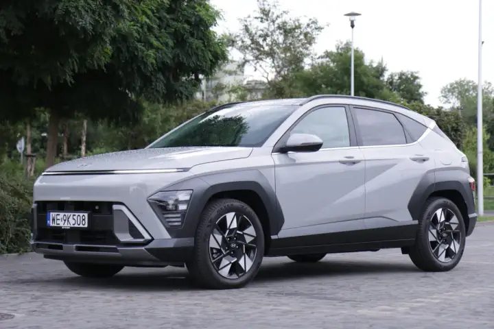 Nowy Hyundai Kona Hybrid w kolorze jasnoszarym, zaparkowany na brukowanej nawierzchni, otoczony zielenią.