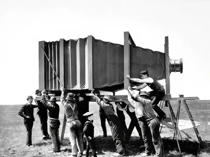 Grupa mężczyzn w historycznych strojach ustawia gigantyczny aparat fotograficzny. Wyobraź sobie, kiedy wynaleziono aparat fotograficzny i jak wielkie musiały być pierwsze urządzenia!
