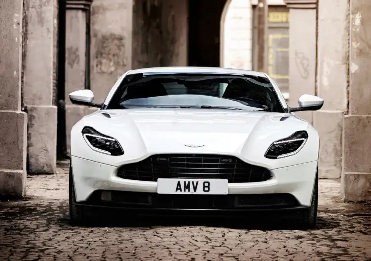 Biały Aston Martin DB11 z tablicą AMV 8 stoi na brukowanej uliczce, otoczony kamiennymi budynkami.