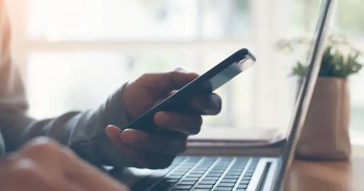 Dłoń trzymająca smartfon nad klawiaturą laptopa.