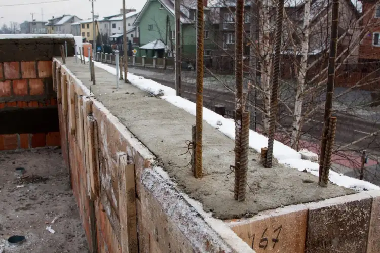 Wylany beton na budowie, a na nim przymrozek. Stalowe pręty wystają z mokrej masy, a wokół leży śnieg.