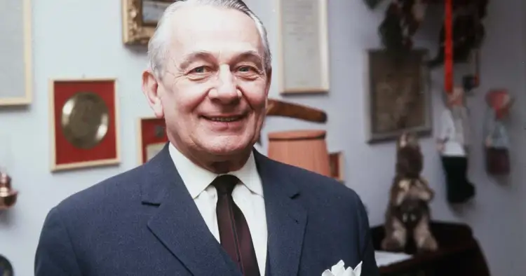 Uśmiechnięty Mieczysław Fogg w garniturze, jakby żył podwójnym życiem, z nagrodami na ścianie.