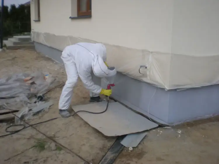 Osoba w kombinezonie maluje cokół budynku metodą natryskową. To pokazuje, jak malować elewację, by uzyskać równomierne krycie.