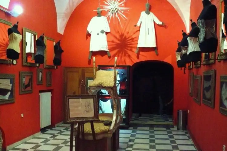 Wnętrze muzeum maszyn erotycznych w Pradze. Czerwone ściany, gorsety na manekinach, wiszące postacie w białych szatach i szachownicowa podłoga.