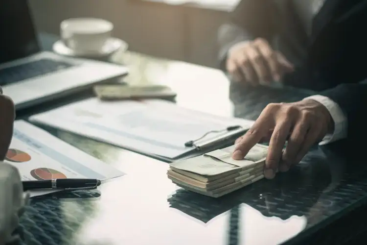 Dłoń sięga po stos banknotów leżący na stole obok laptopa i dokumentów.