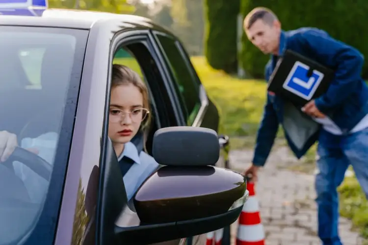 Dziewczyna za kierownicą, zdenerwowana, czeka na egzaminatora z teczką "L". Tak wygląda egzamin na prawo jazdy.