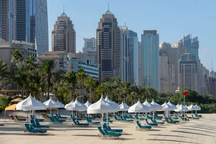 Piękna dubaj pogoda luty: pusta plaża z leżakami i białymi parasolami, w tle palmy i nowoczesne wieżowce.