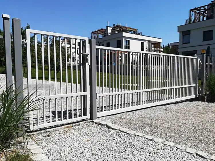 Brama przesuwna na pilota, biała, z pionowymi szczebelkami, otwiera wjazd na posesję z nowoczesnymi budynkami.