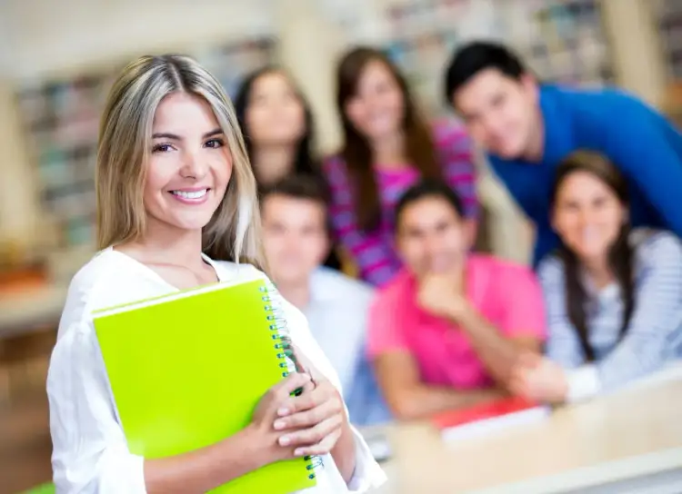 Uśmiechnięta studentka z zielonym zeszytem, gotowa do rozwiązywania zadań z biologii. W tle grupa innych studentów.