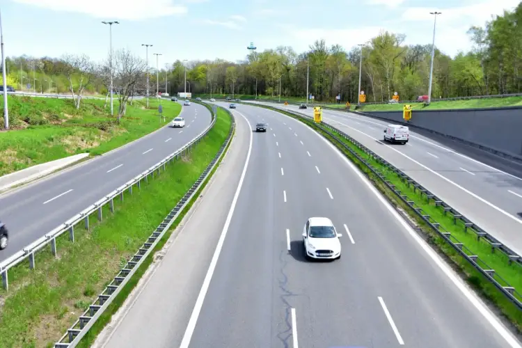 Autostrada z zielonymi pasami i drzewami. Samochody jadą z różną prędkością, ale żadne nie przekracza minimalnej prędkości na autostradzie.