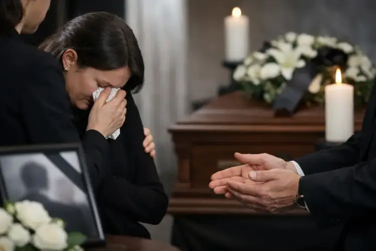 Smutna kobieta ociera łzy chusteczką, wspierana przez bliską osobę. W tle trumna, kwiaty i świece. Słowa pocieszenia na pogrzebie rodzinie.