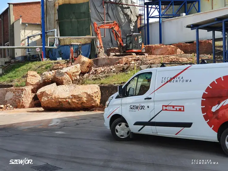 Wyburzenie ceglanych bloków i pytanie: ile kosztuje kucie betonu? Na placu budowy koparka i biały van z napisem "Technika Diamentowa".