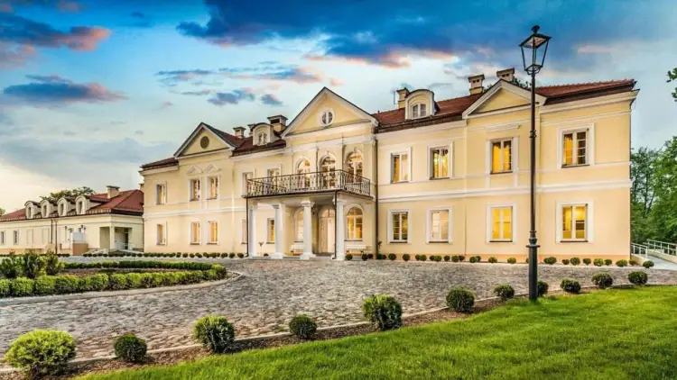 Elegancki hotel Sobienie Królewskie w żółtej barwie, z brukowanym podjazdem i zadbanym ogrodem, pod błękitnym niebem.