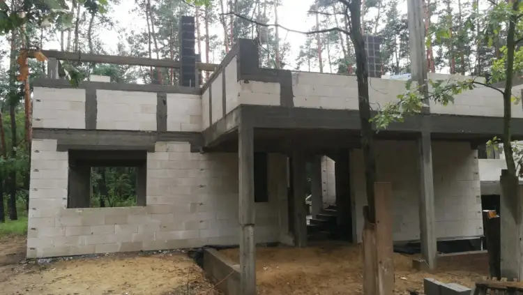 Budowa domu w lesie. Widoczne białe bloczki, betonowe słupy i trzpienie żelbetowe w ścianach murowanych.