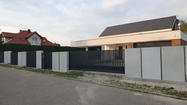 Nowoczesne ogrodzenie z beton architektoniczny i ciemnoszarymi panelami, otaczające posesję z domem.